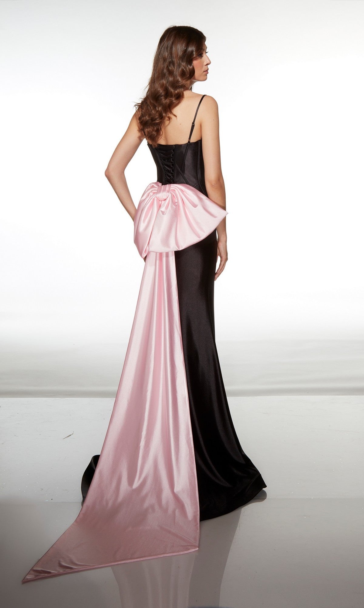 A woman wears the Long Prom Dress: Alyce 61983, featuring black stretch satin, thin straps, a corset bodice, a large pink bow, and a dramatic detachable sweep train. The look is set against a white background.